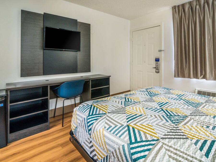 A hotel room with a bed, a desk with a chair, a television mounted on the wall, a door, and a window with curtains.