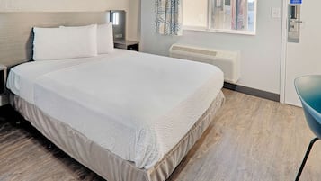 Un lit bien fait, recouvert d’une couvre-lit blanche et agrémenté d’oreillers, une table de chevet avec une lampe, une fenêtre donnant sur un camion Coca-Cola, et un climatiseur fixé au mur.