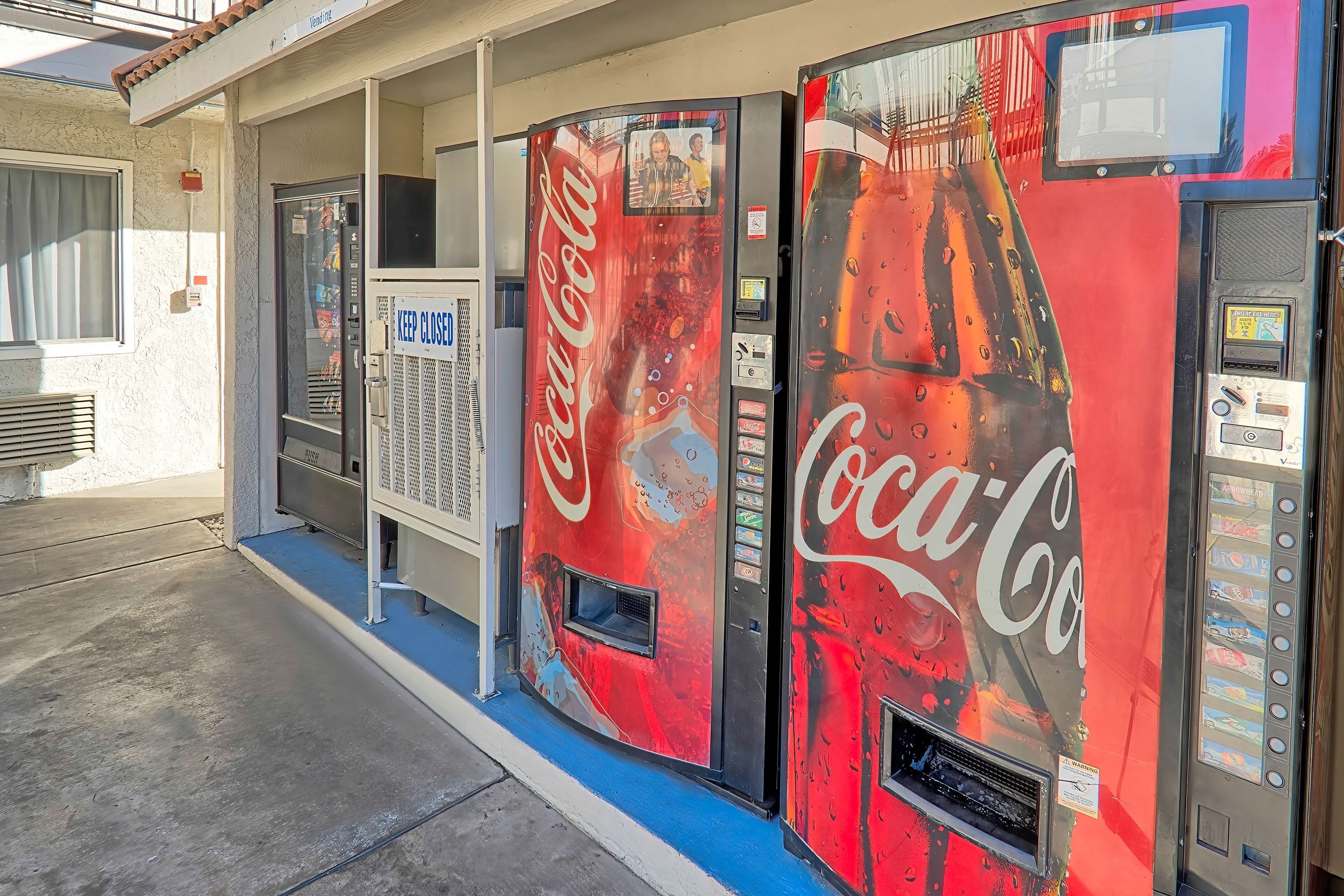 Máquina automática de Coca-Cola com interface de pagamento digital, situada fora de um edifício com toldo azul.