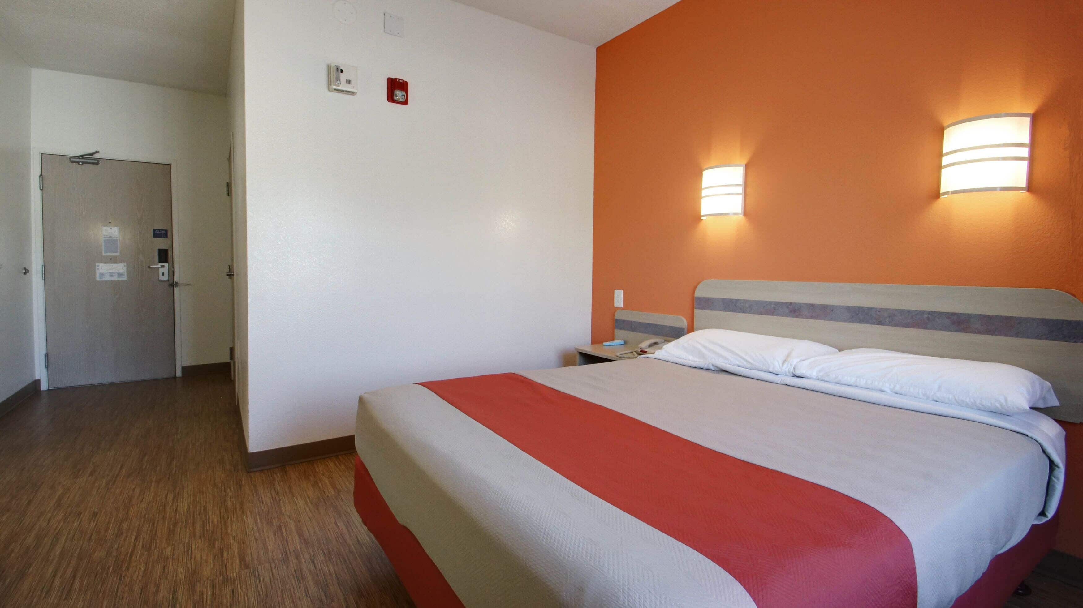 A hotel room with a bed, a nightstand, a wall-mounted lamp, and a door.