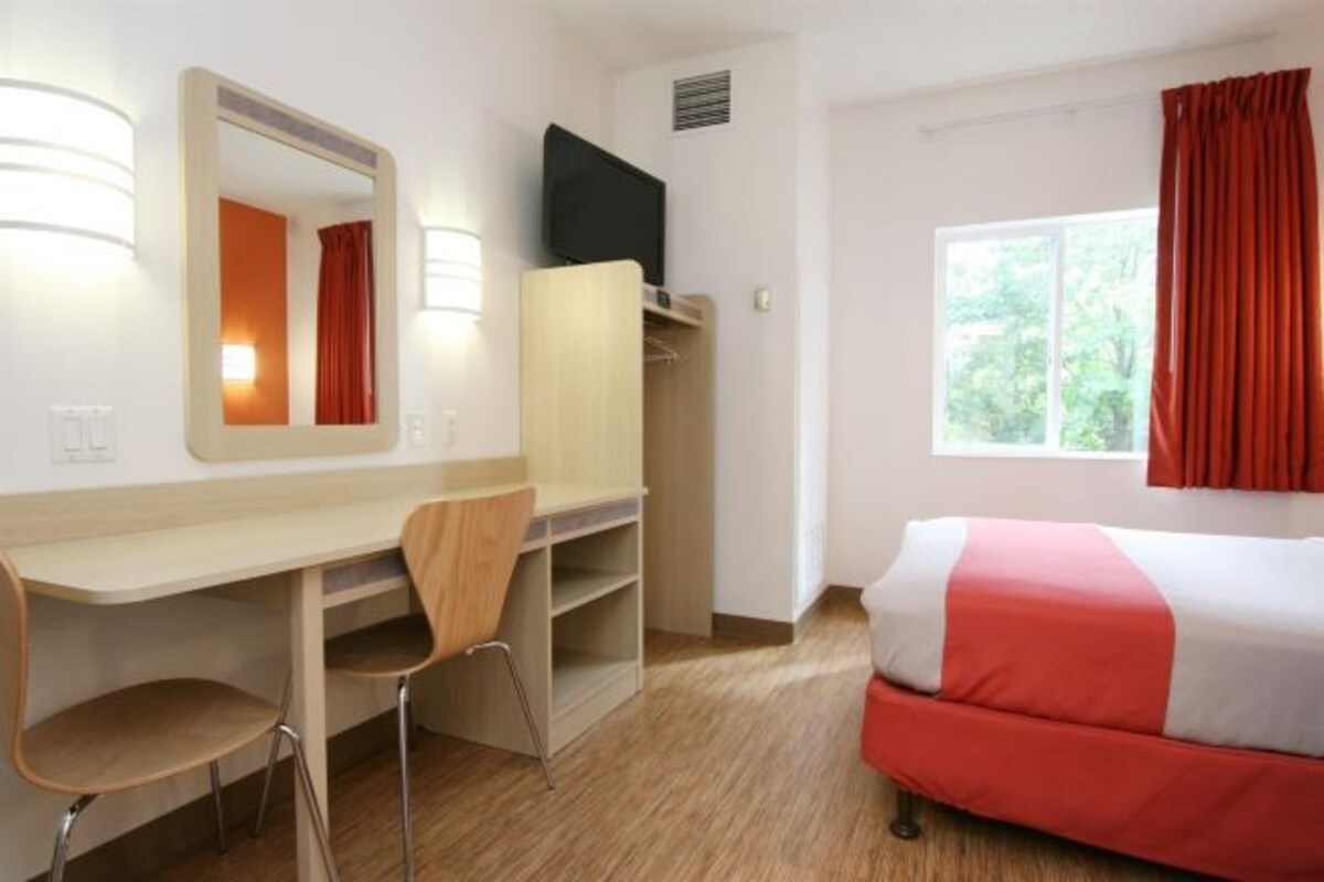 A hotel room with a bed, a desk with a chair, a mirror, a television, and a window with curtains.