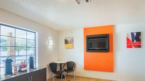 A modern room with a large window, a small table with two chairs, a TV mounted on an orange wall, and a cabinet with a coffee machine and cups. - Motel 6 Lawton, OK (Lawton)