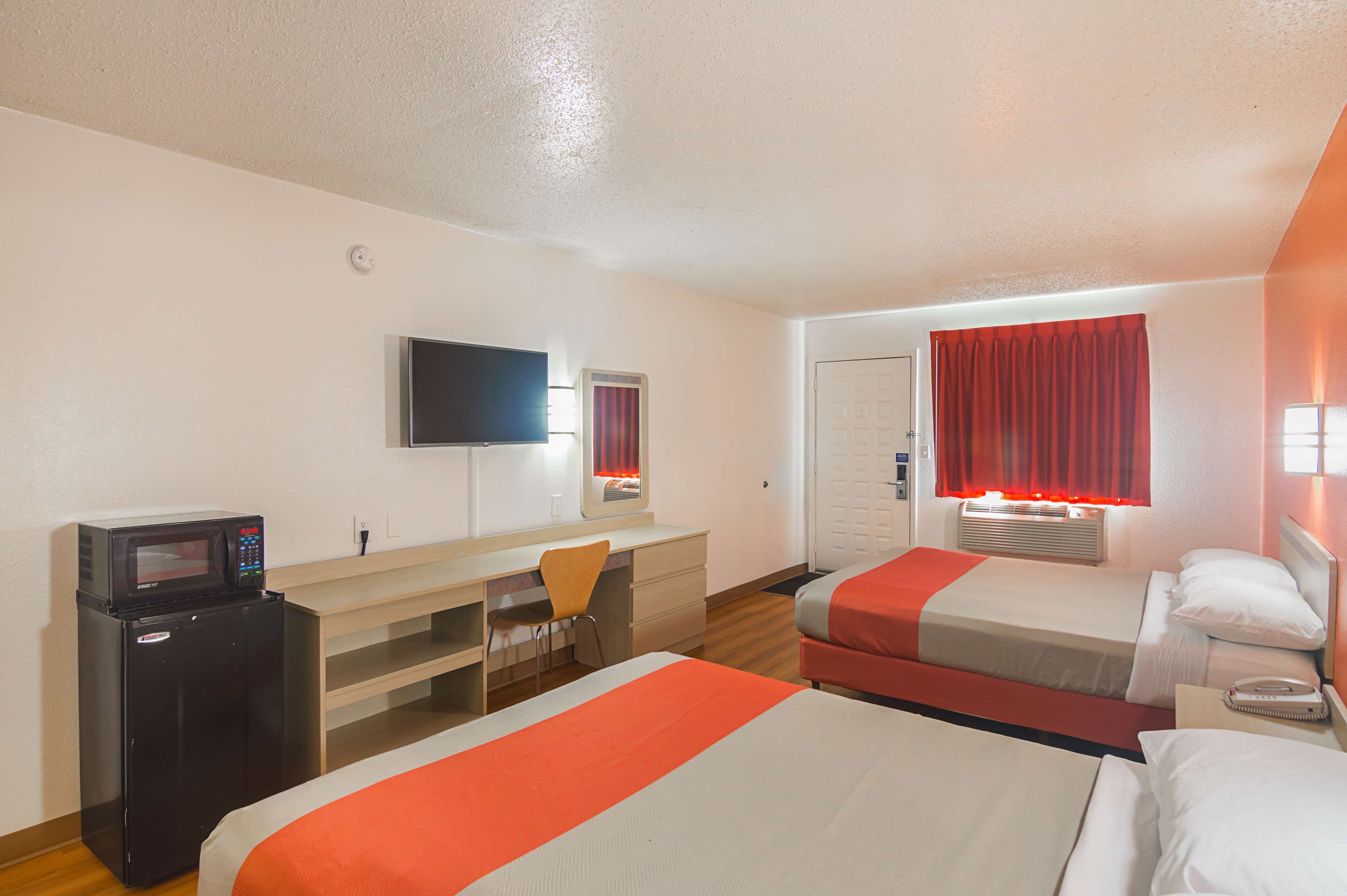 Habitación de hotel con dos camas, un escritorio con silla, microondas, televisor y ventana con cortinas rojas.