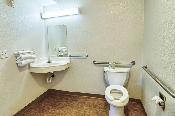 Une salle de bain avec un lavabo blanc, un miroir et des toilettes. Des serviettes sont pliées et empilées sur un support mural, il y a une barre d’appui et un porte-rouleau de papier toilette.