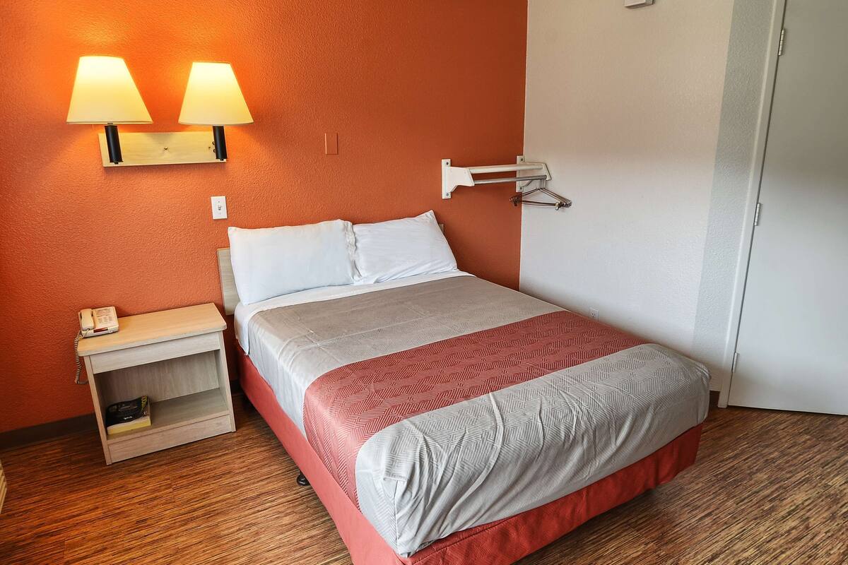 A hotel room with a bed, bedside lamp, nightstand, and a wall-mounted light fixture.
