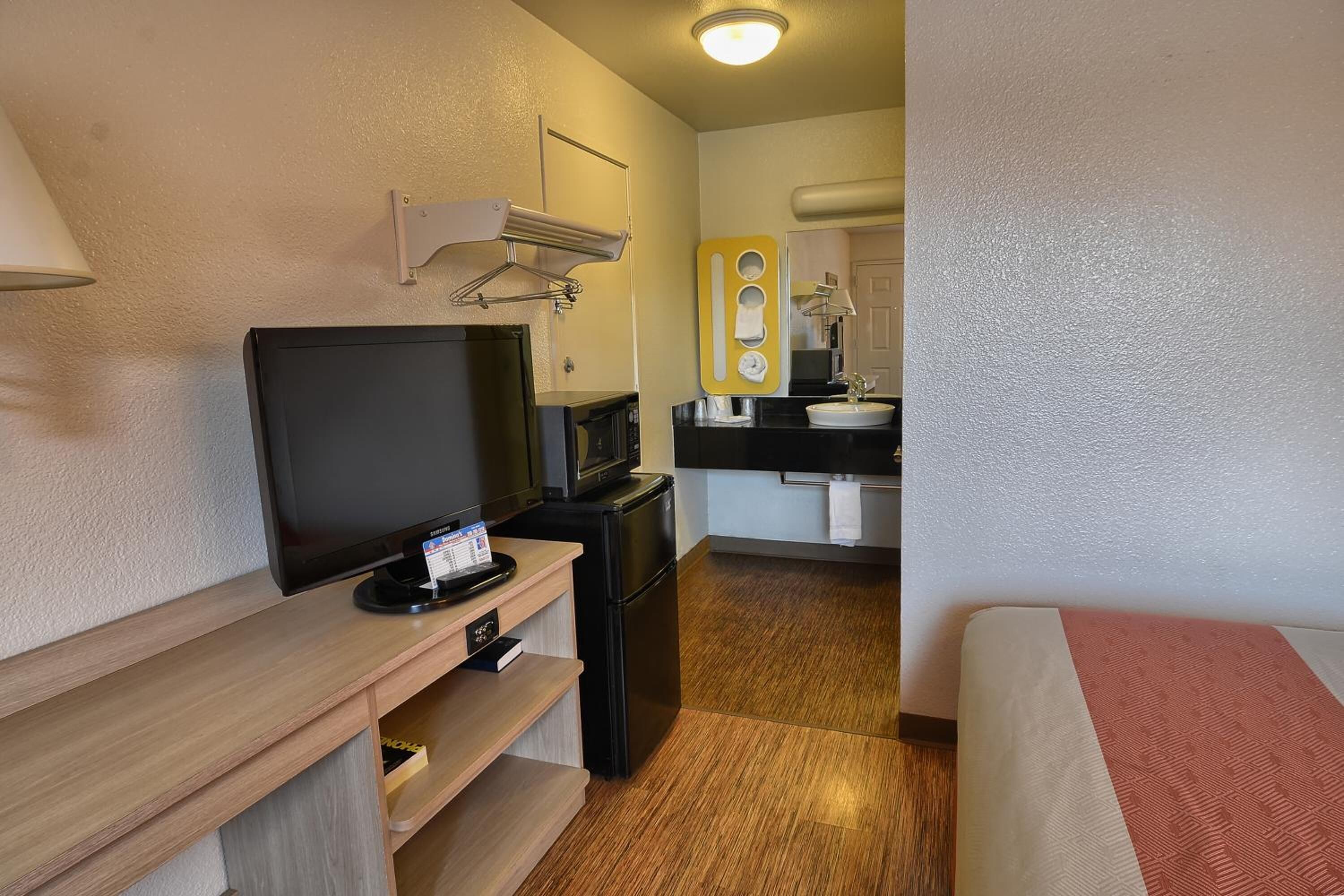A compact hotel room with a wooden desk, a television, a microwave, a small refrigerator, a bed, and a bathroom with a sink and mirror.