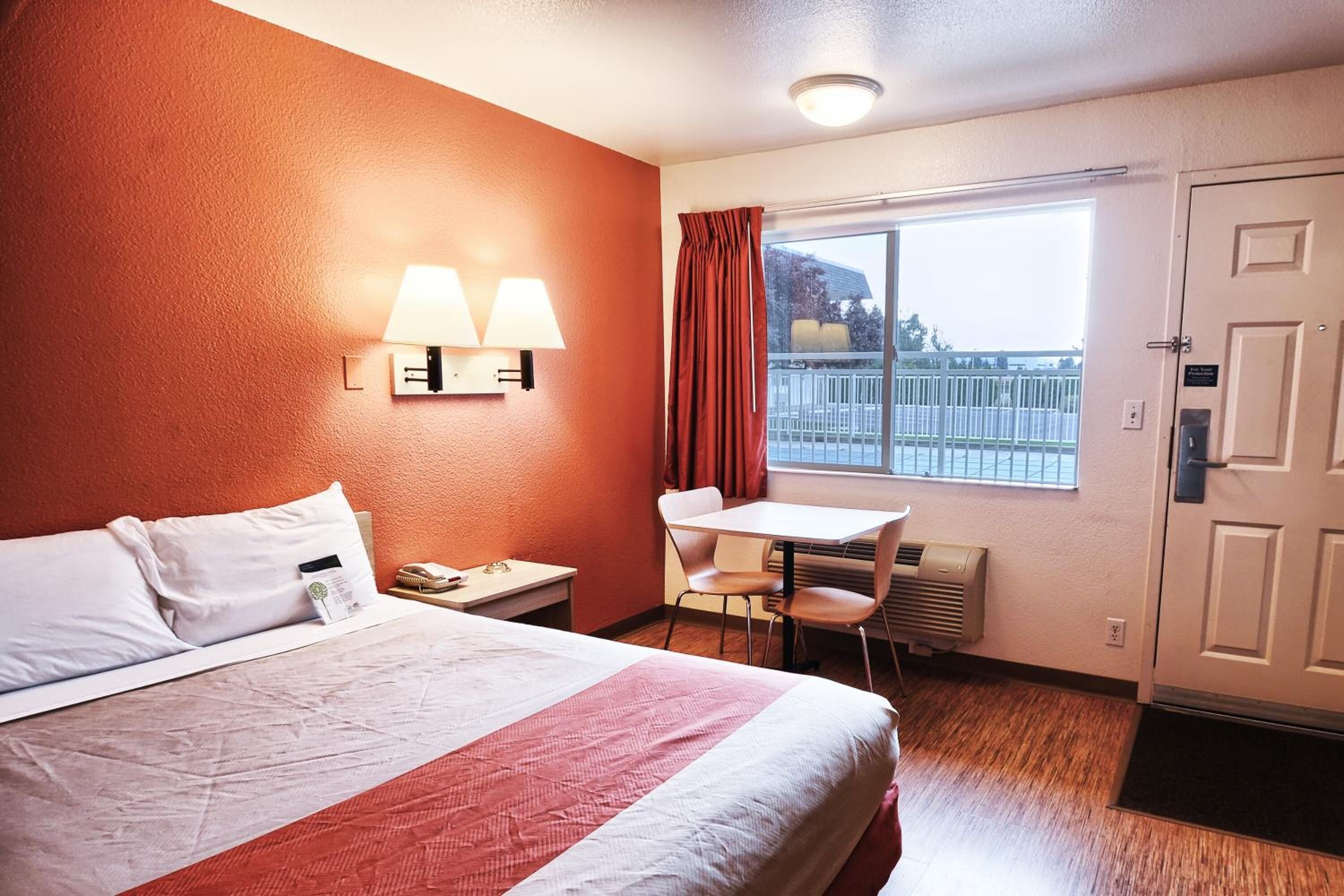 A hotel room with a bed, a desk, two wall-mounted lamps, a window with curtains, and a door.