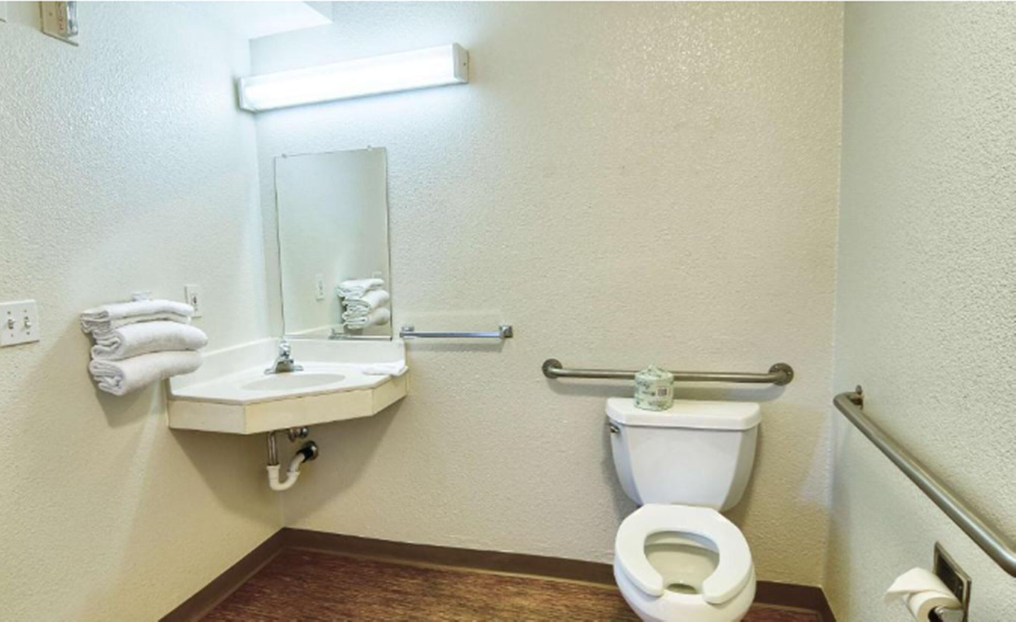 A small, clean bathroom with a sink, mirror, toilet, and handrails.