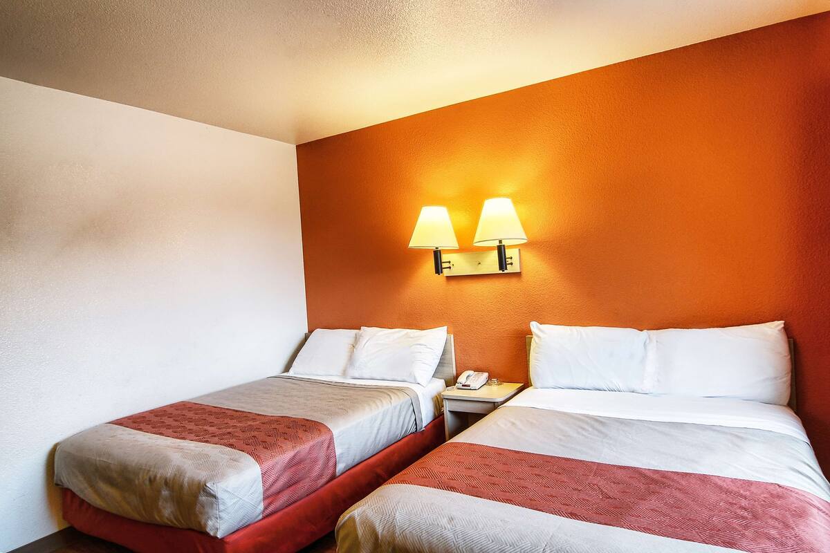 A hotel room with two beds, each with a red and white bedspread, and a wall-mounted lamp above the beds.