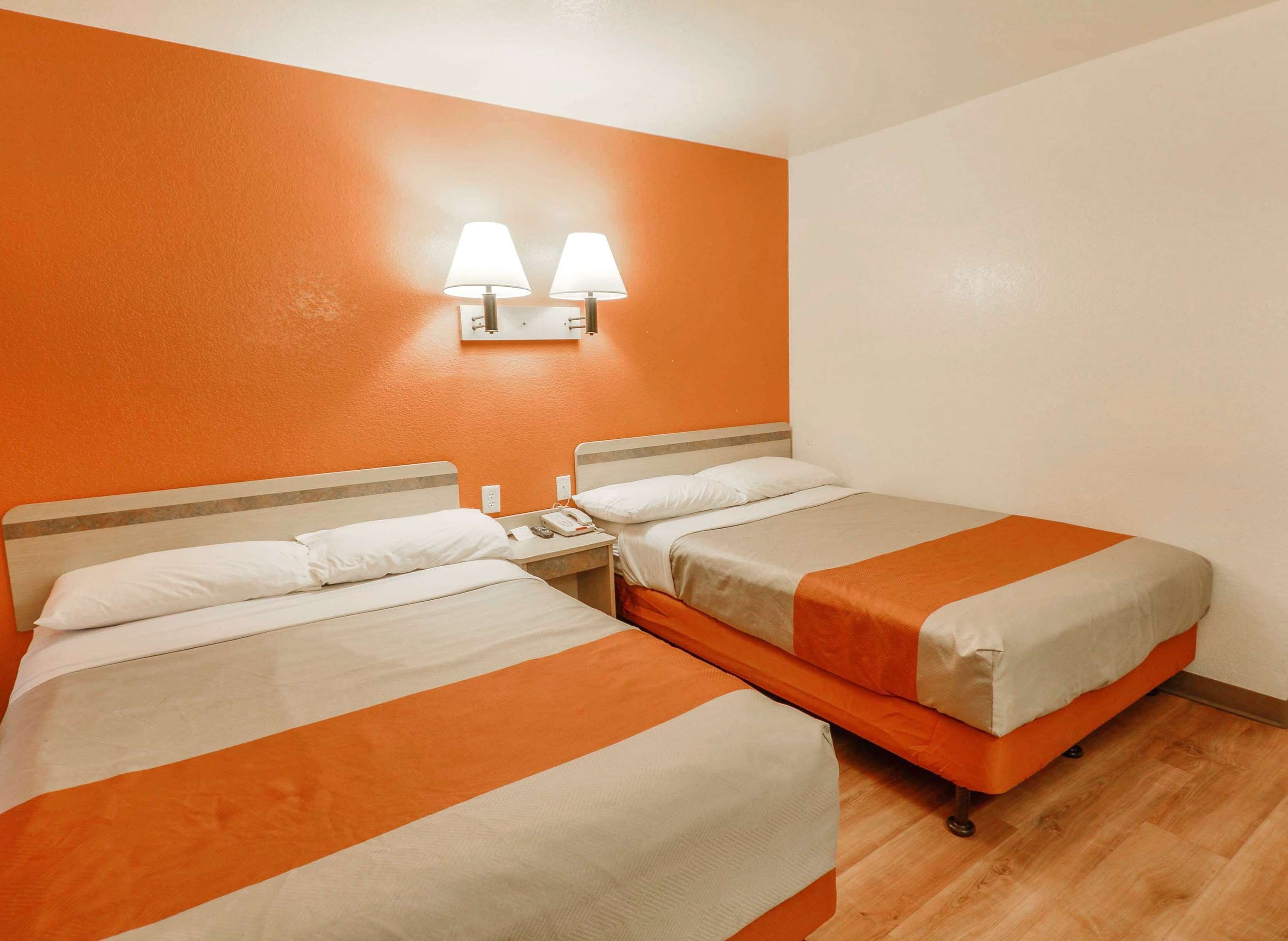 A hotel room with two beds, each with white and orange bedding, a nightstand with a phone and lamp, and an orange accent wall with two wall-mounted lamps.