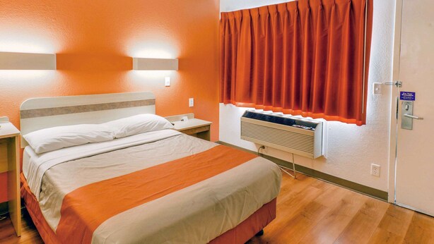 A hotel room with a bed, a nightstand, a window with orange curtains, a wall-mounted air conditioner, and a door with a lock. - Motel 6 Huntsville, TX (Huntsville)