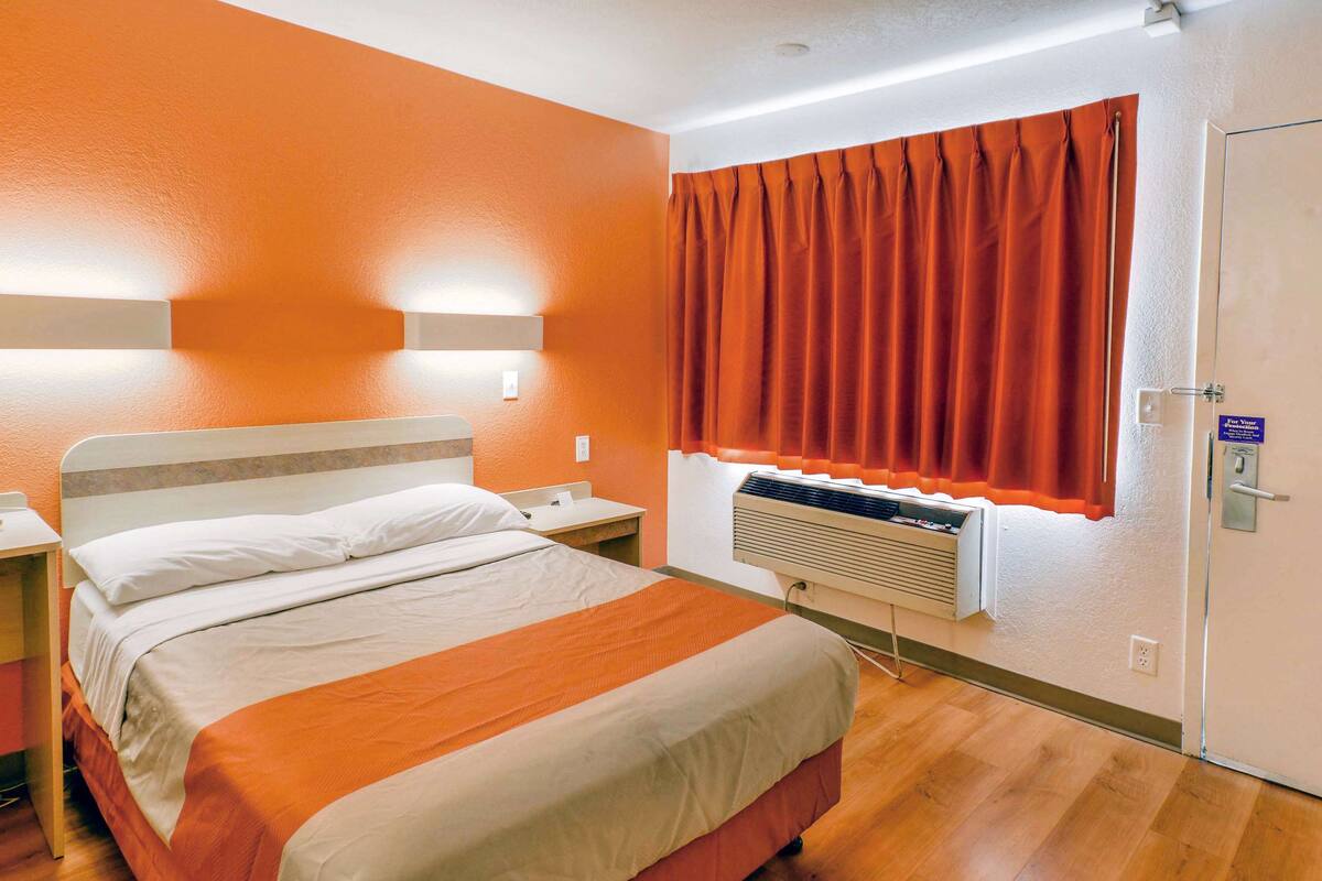 A hotel room with a bed, a nightstand, a window with orange curtains, a wall-mounted air conditioner, and a door with a lock.