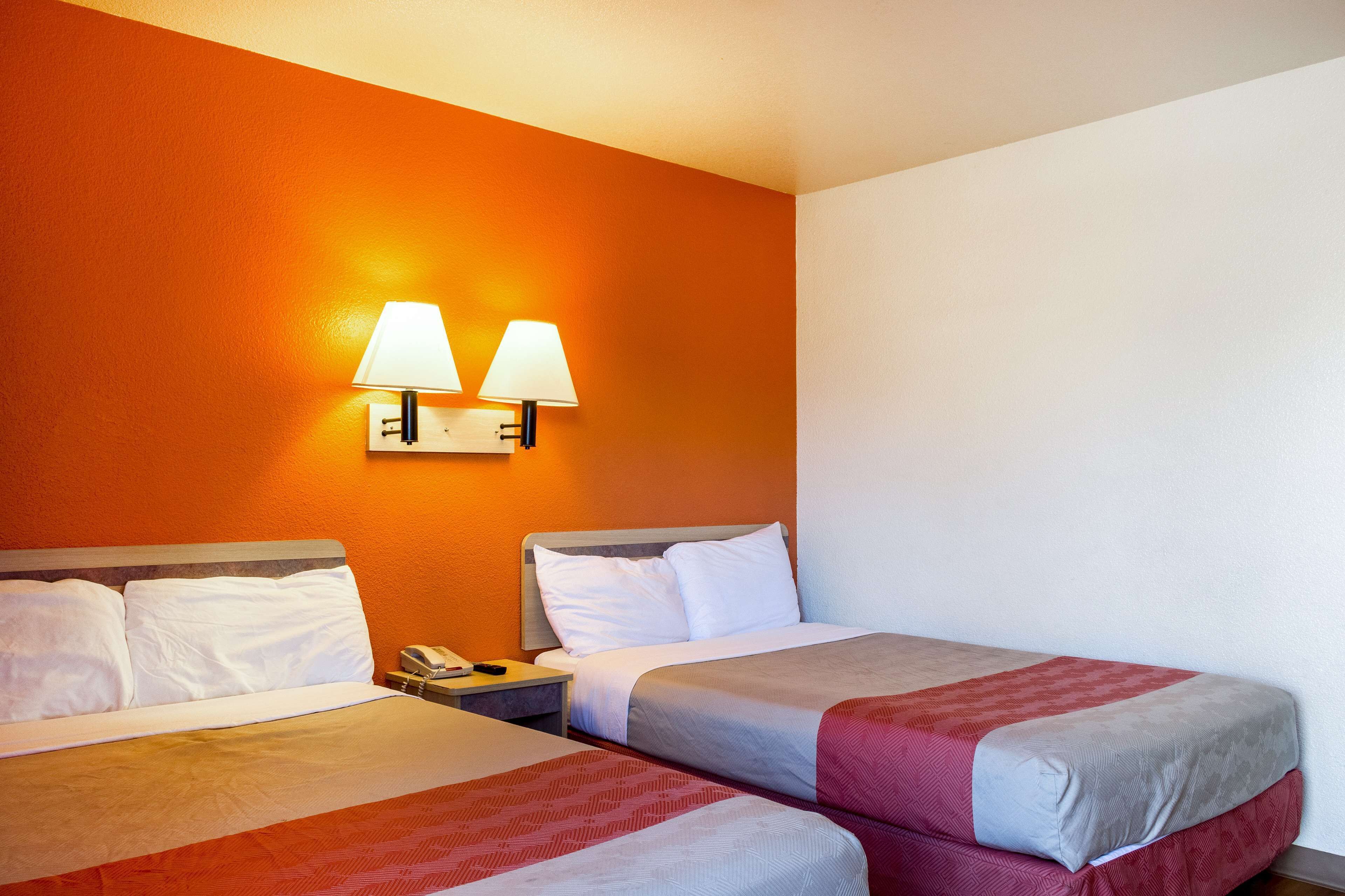 A hotel room with two beds, each with white linens and a red and grey bedspread. There are two wall-mounted lamps on the orange wall.