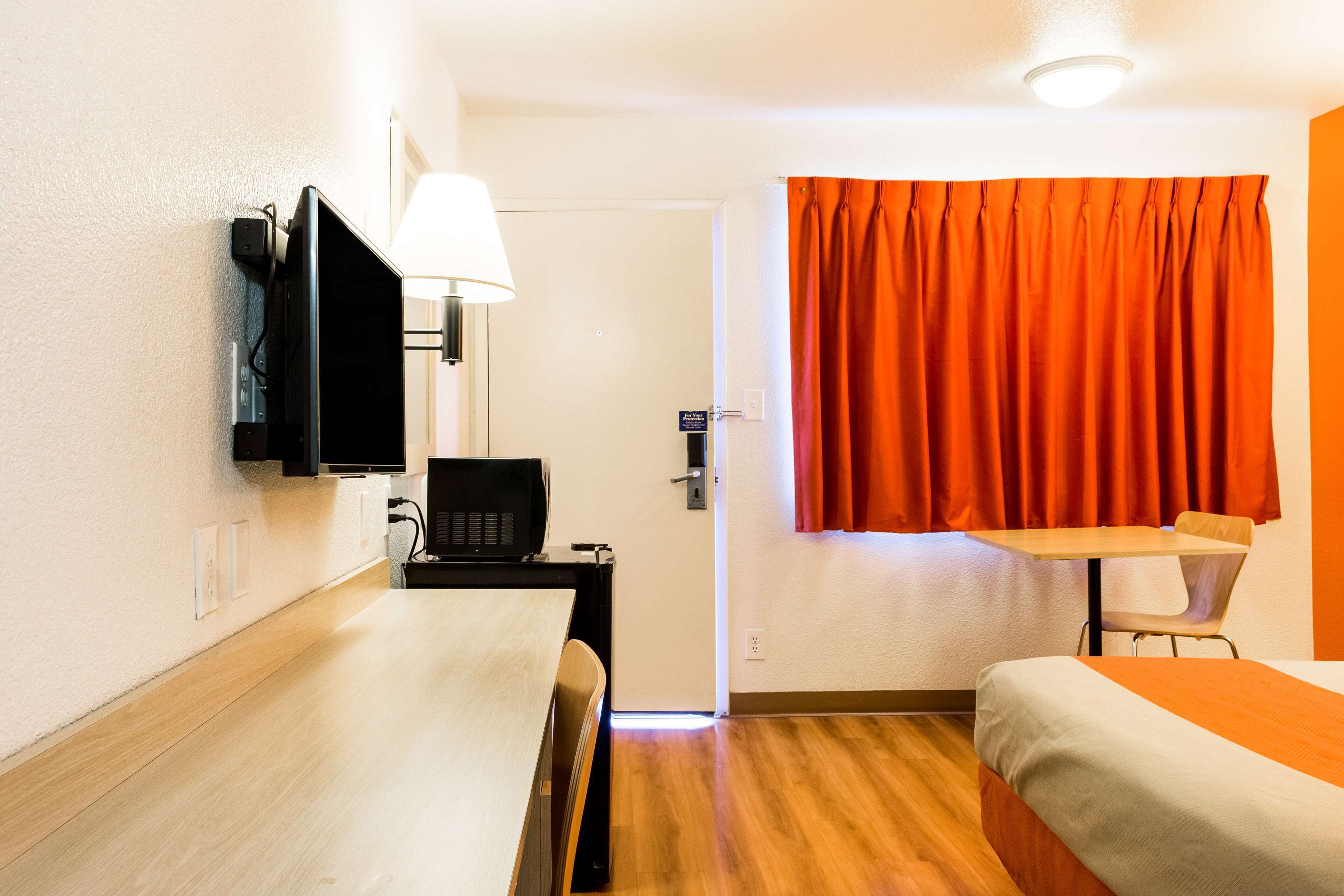 Hotellihuone, jossa on sänky, työpöytä tuolilla, televisio ja ikkuna, jossa on oranssit verhot.