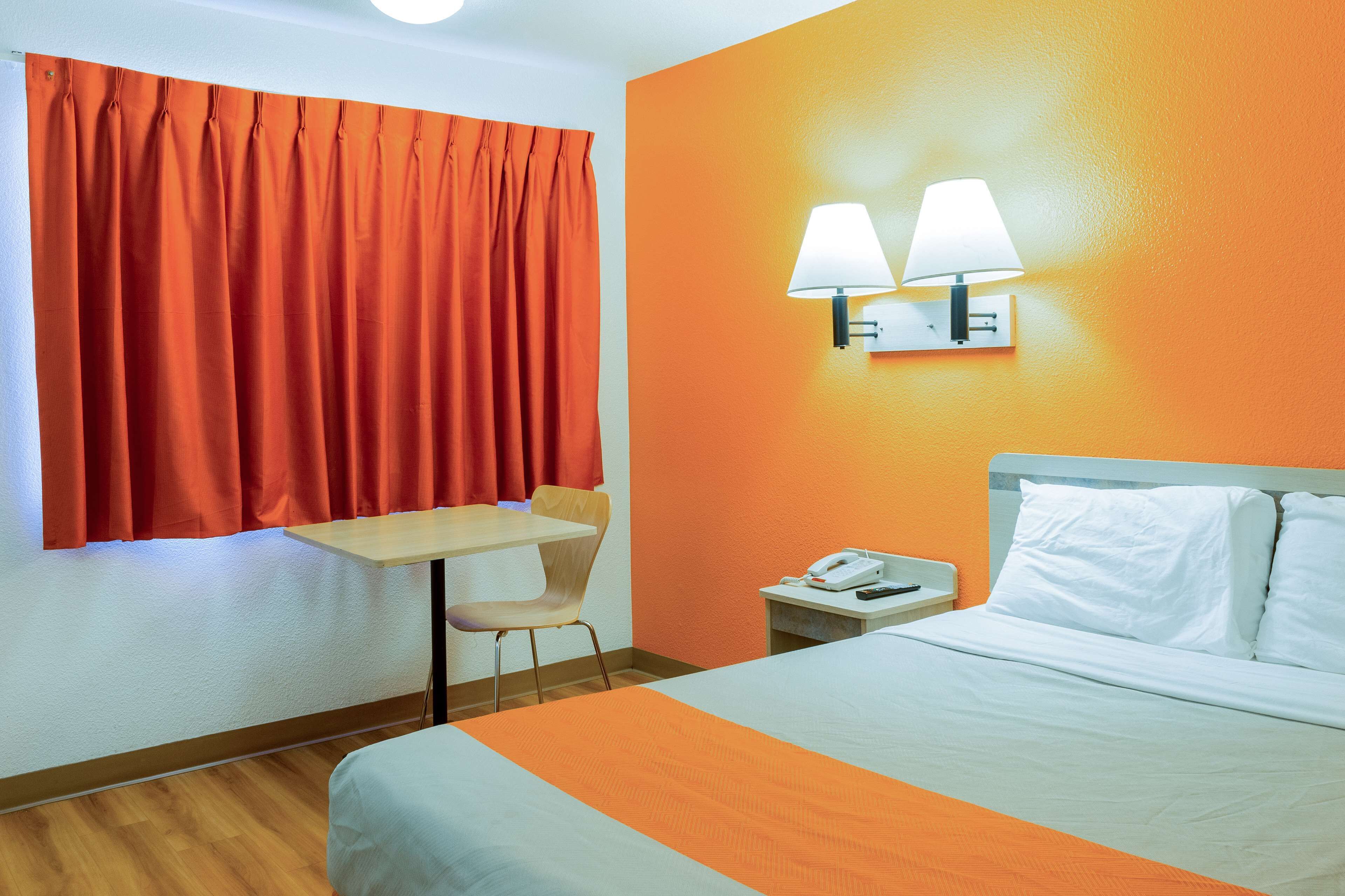 A hotel room with a bed, a desk, a chair, a nightstand with a phone, and two wall-mounted lamps.