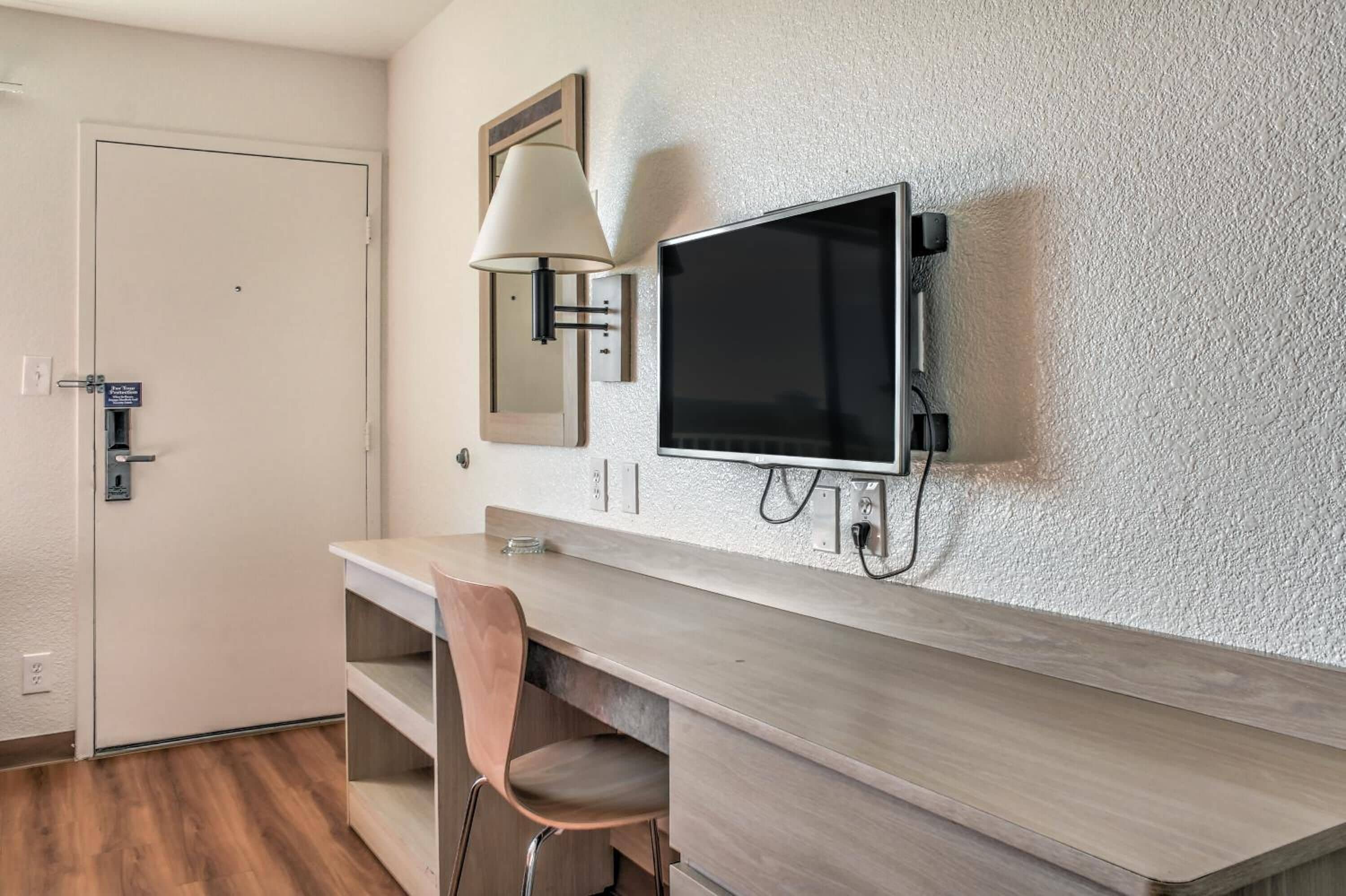 Una habitación de hotel moderna con un escritorio de madera, una silla, un televisor de pantalla plana montado en la pared y una puerta con lector de tarjeta.
