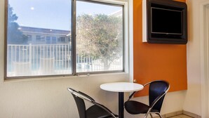 A room with a large window, a small round table, two black chairs, and a mounted television on the wall. - Motel 6 Mojave, CA (Mojave)