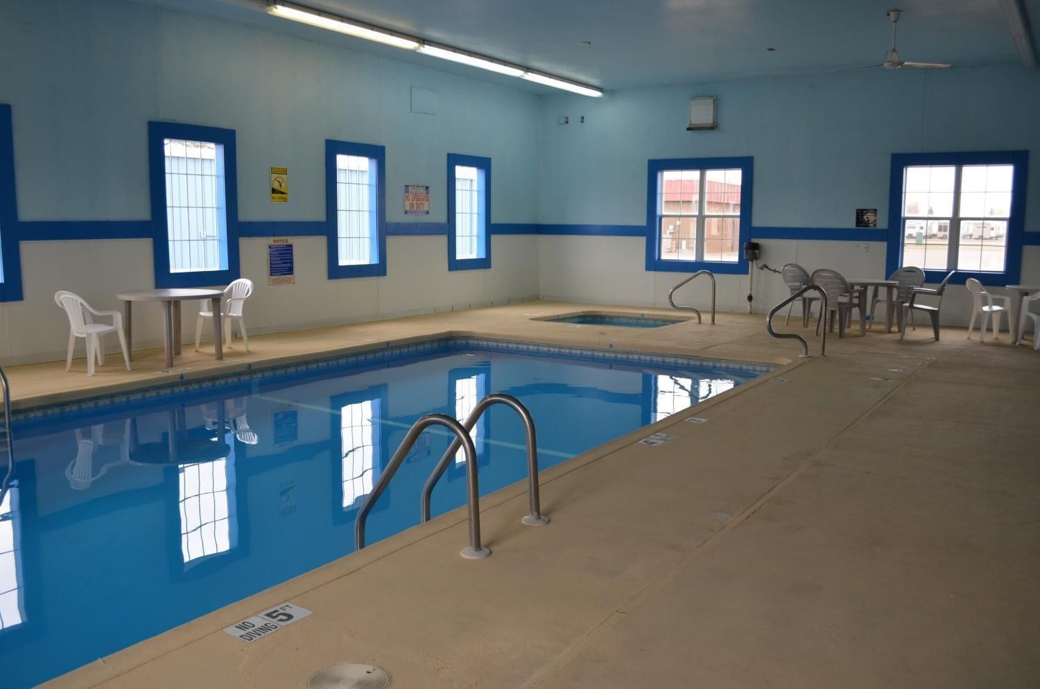 An indoor swimming pool with clear blue water, surrounded by white plastic chairs and tables.