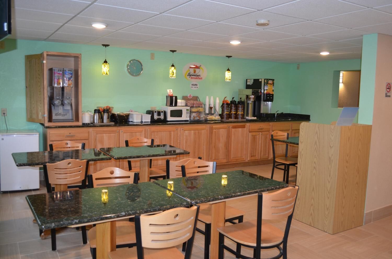 A small, cozy restaurant with wooden furniture and a green wall. There are several tables and chairs, a refrigerator, and a microwave on the counter.