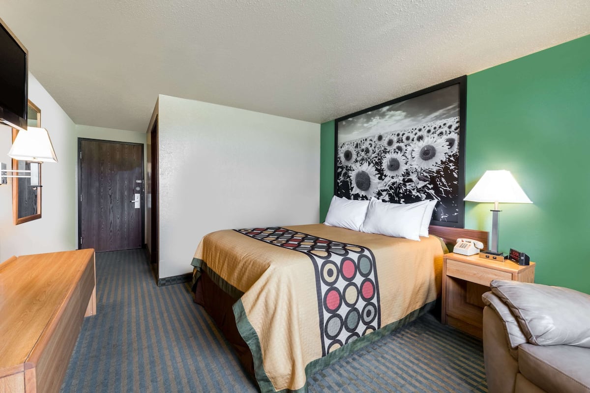 A hotel room with a bed, a desk, a chair, a nightstand with a lamp, and a painting on the wall.