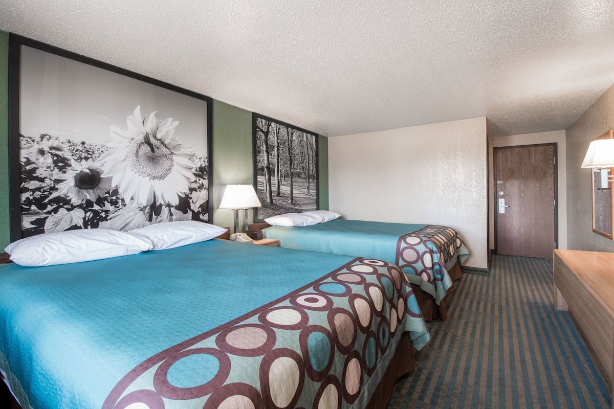 A hotel room with two beds, a large floral artwork on the wall, a patterned carpet, and a wooden door.