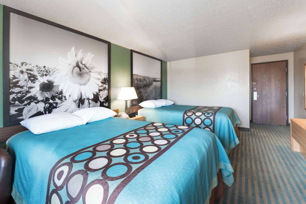 A hotel room with two beds, a large floral artwork on the wall, a bedside lamp, and a wooden door.
