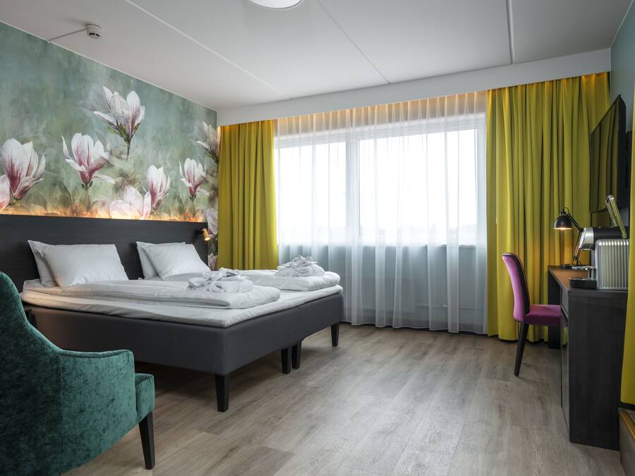 Quarto de hotel moderno com cama, escrivaninha, cadeira e uma janela grande com cortinas amarelas.