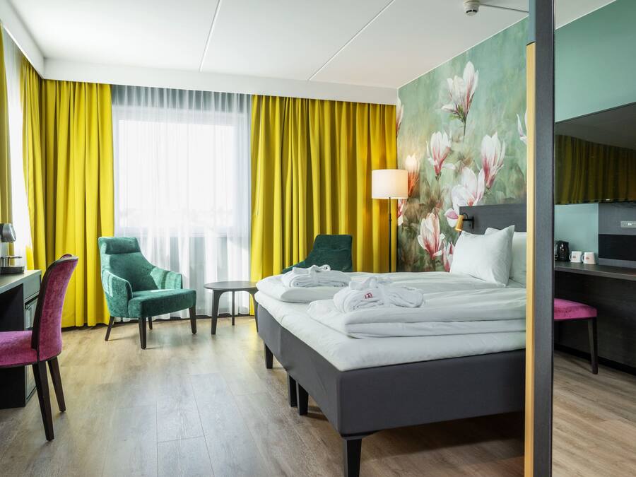 A hotel room with a bed, a desk, a chair, a small table, and a lamp.