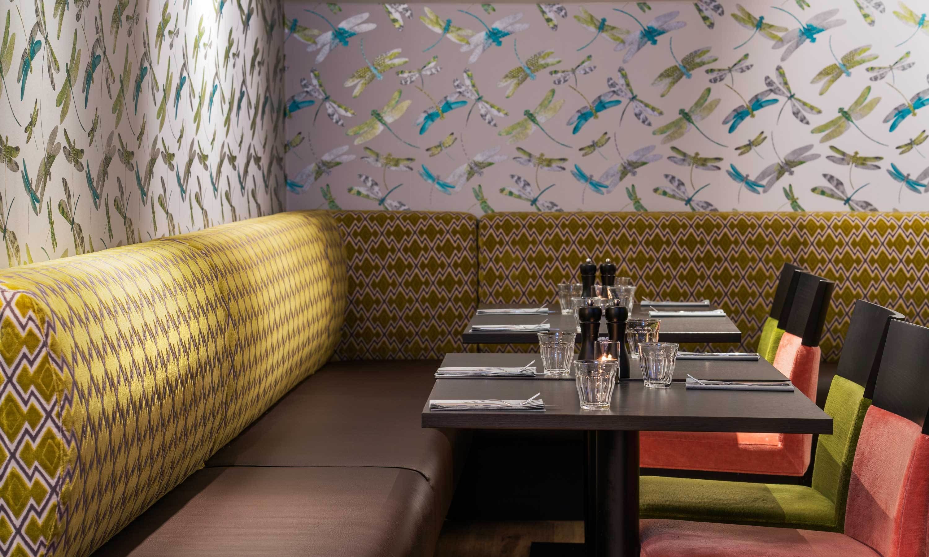 A restaurant interior with a patterned wall, a cushioned booth, and a table set with glasses and cutlery.