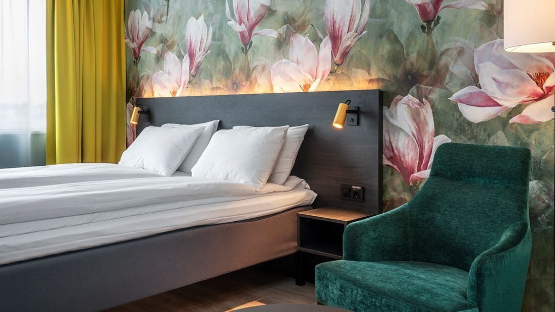 A bed with white linens, a green armchair, and a wall with a floral mural.