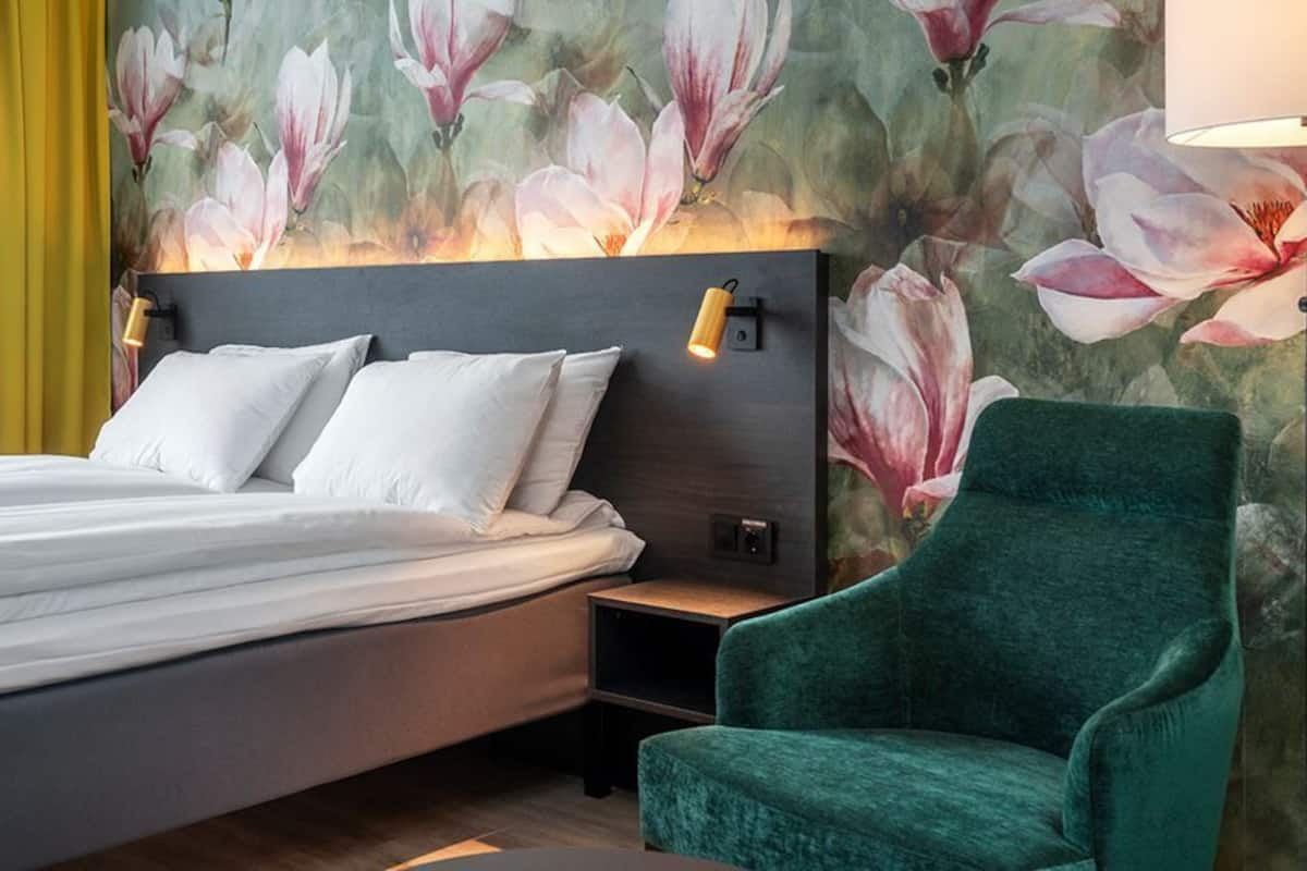 Una cama con sábanas blancas, un sillón verde y una pared con un mural floral.