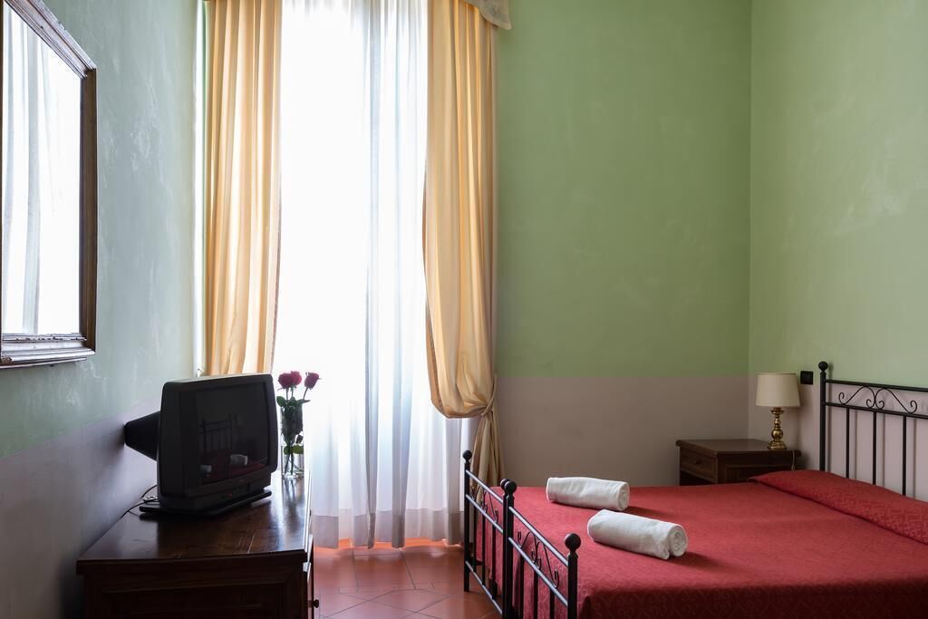 Ein Hotelzimmer mit einem Bett, einem Schreibtisch mit Computer, einer Lampe und einem Fenster mit Vorhängen.