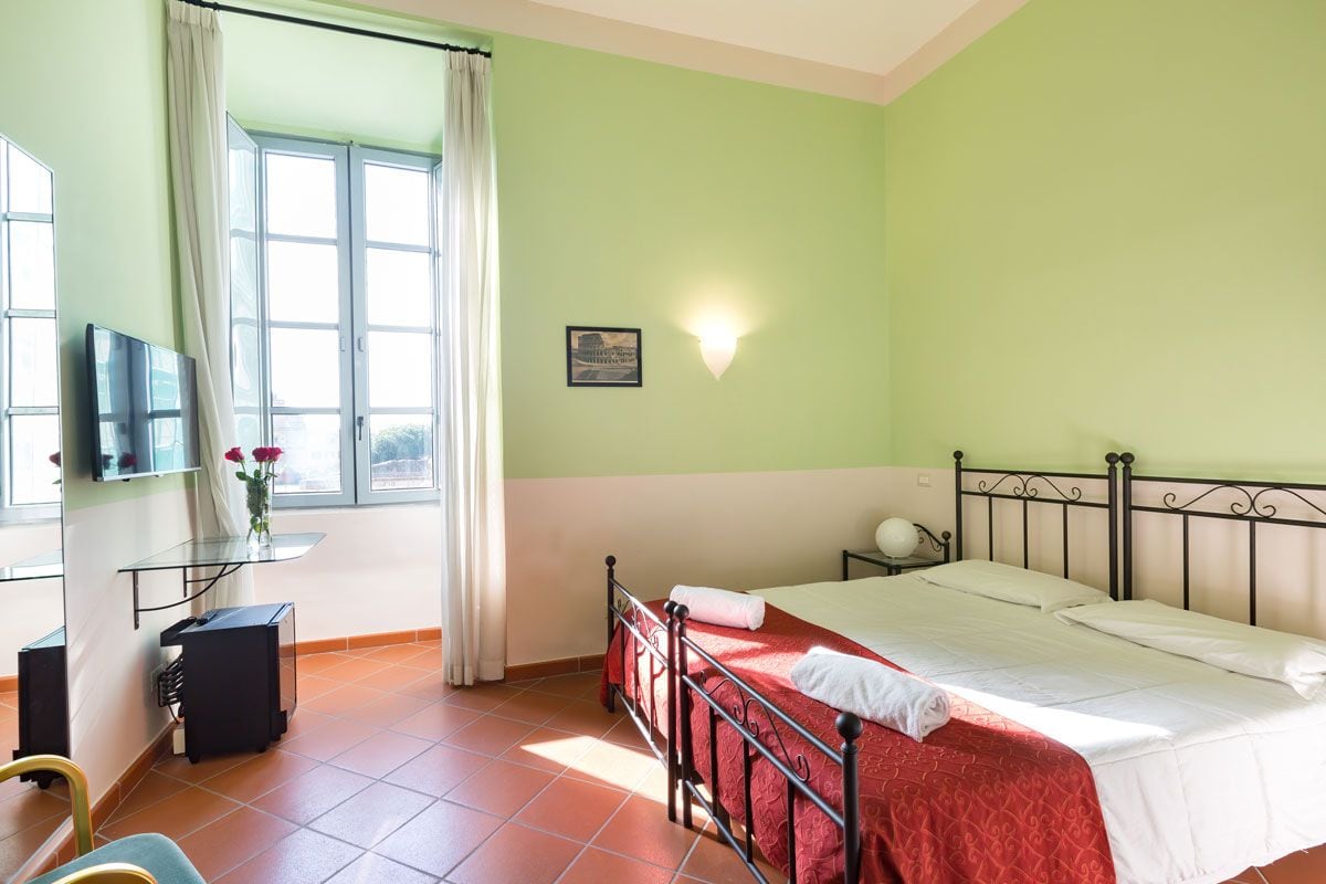 A room with a bed, a desk with a computer, a chair, a window with curtains, and a wall-mounted light.