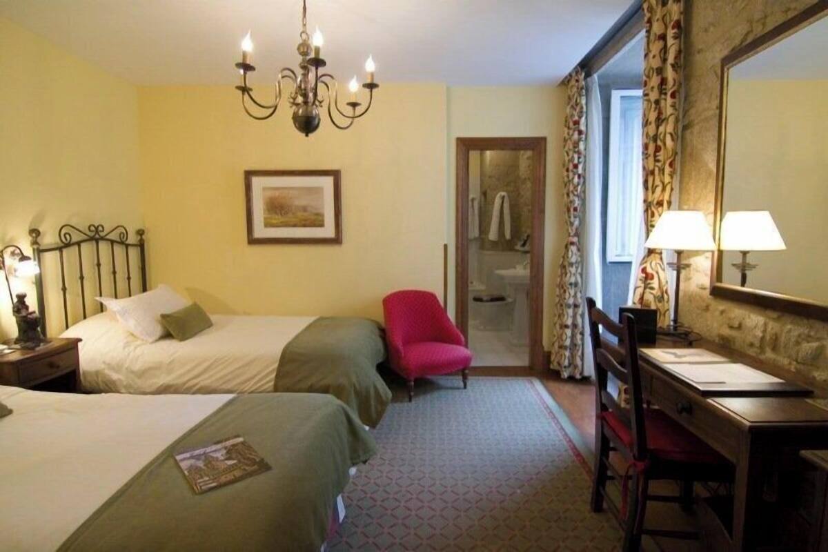 Une chambre d’hôtel avec deux lits, un bureau, une chaise, un miroir, un lustre et une salle de bain visible à travers une porte.