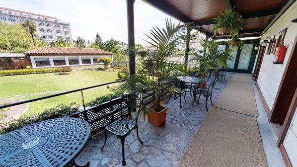 A covered outdoor area with stone flooring, metal tables and chairs, potted plants, and a view of a building in the background. - Hotel Virxe da Cerca by Pousadas de Compostela (Santiago de Compostela)