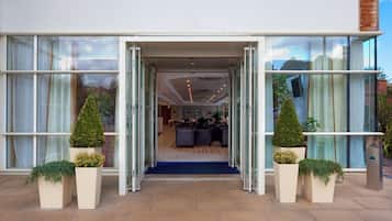 A hotel entrance with a glass facade, a blue carpet leading inside, and potted plants on either side.