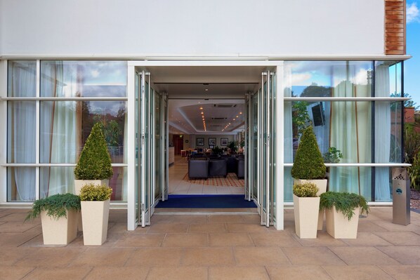 A hotel entrance with a glass facade, a blue carpet leading inside, and potted plants on either side. - Holiday Inn Express Chester - Racecourse by IHG (Chester)