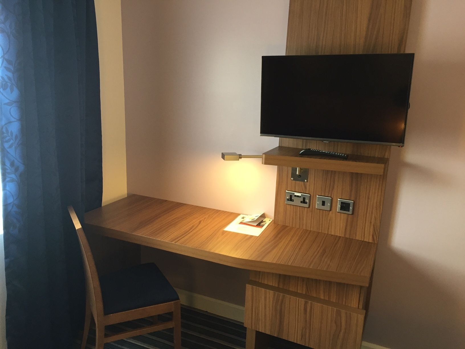 A wooden desk with a television mounted above it, a lamp, and a chair.