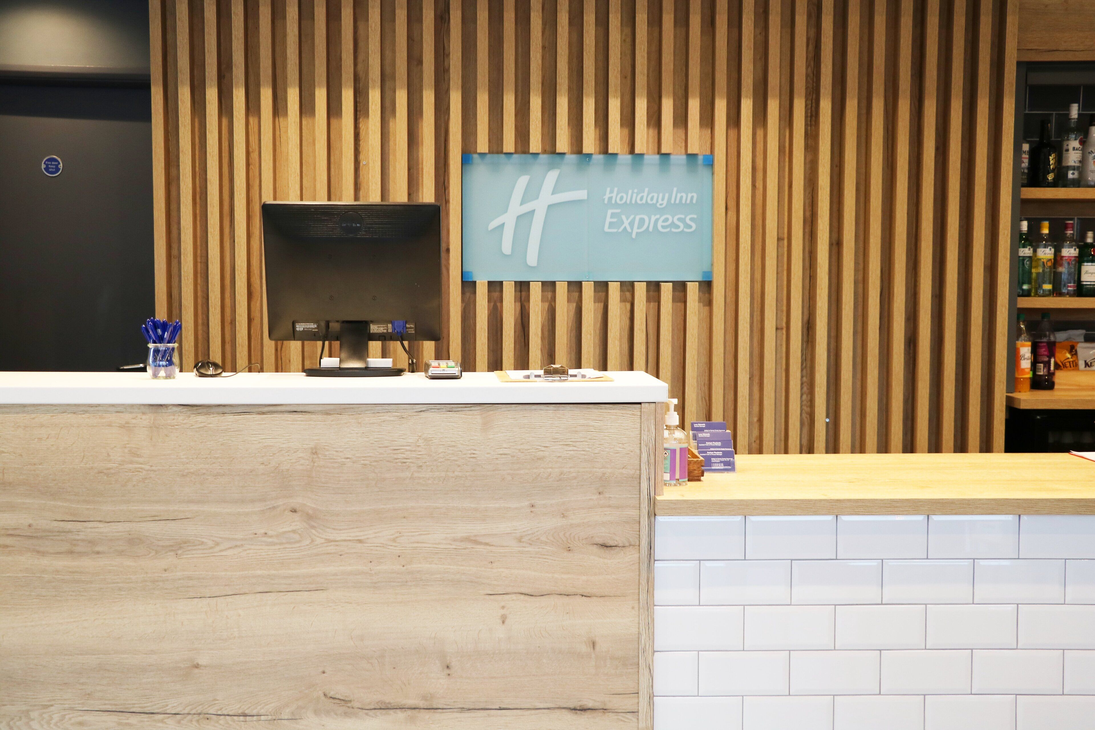 A Holiday Inn Express reception desk with a wooden panel wall, a digital screen, and a counter with amenities.
