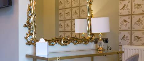 A room with a vanity table, a mirror with an ornate gold frame, a chair, and a lamp.