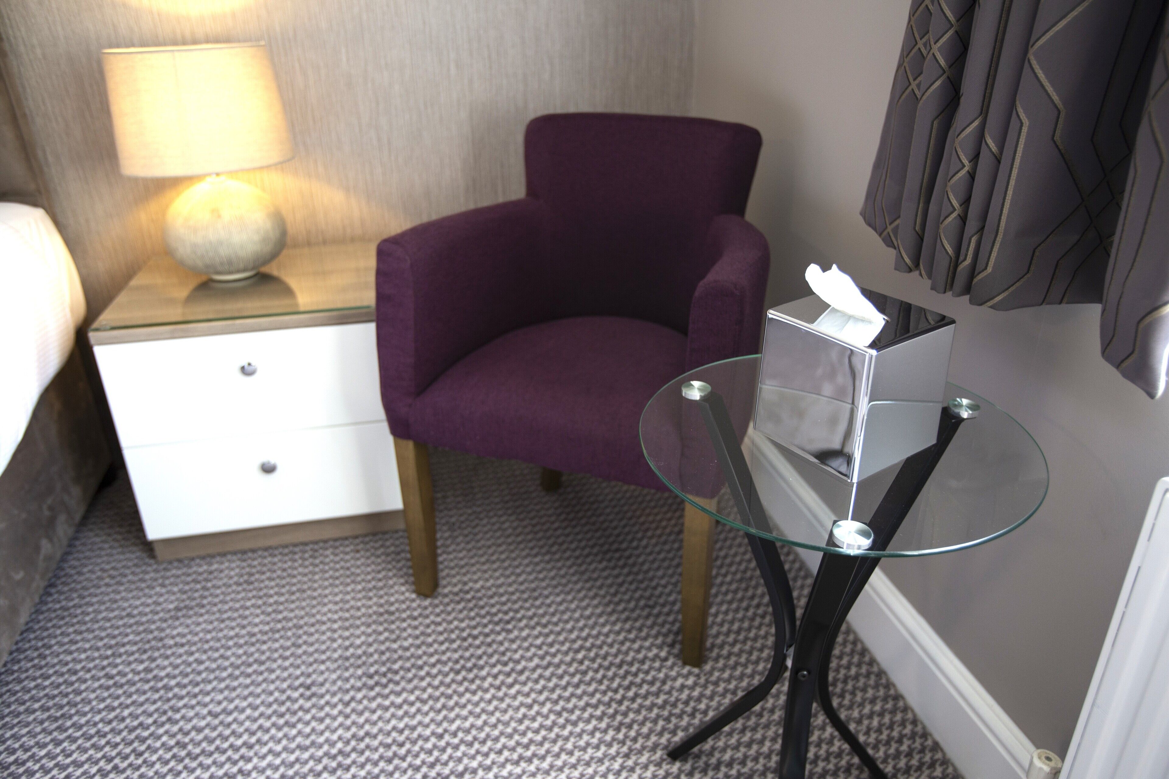 Un fauteuil violet, une table en verre, une lampe et une table de chevet avec une boîte de mouchoirs, dans une chambre d’hôtel.