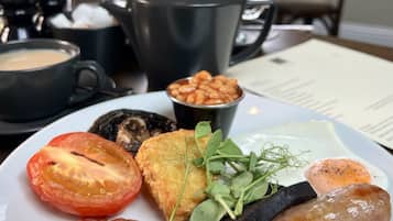 Un plato de desayuno con tocino, salchicha, tomate, champiñones, frijoles y vegetales de hoja verde. Se ven dos vasos de jugo de naranja y una tetera al fondo.