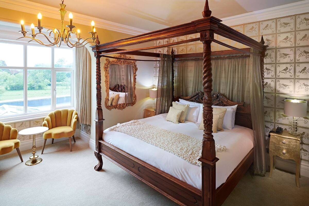 A four-poster canopy bed with a wooden frame, a round table with a gold base, and two armchairs.