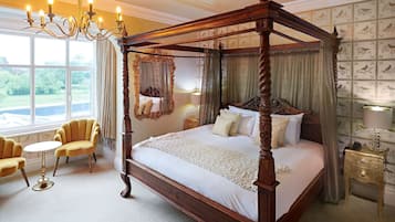 A four-poster canopy bed with a wooden frame, a round table with a gold base, and two armchairs.