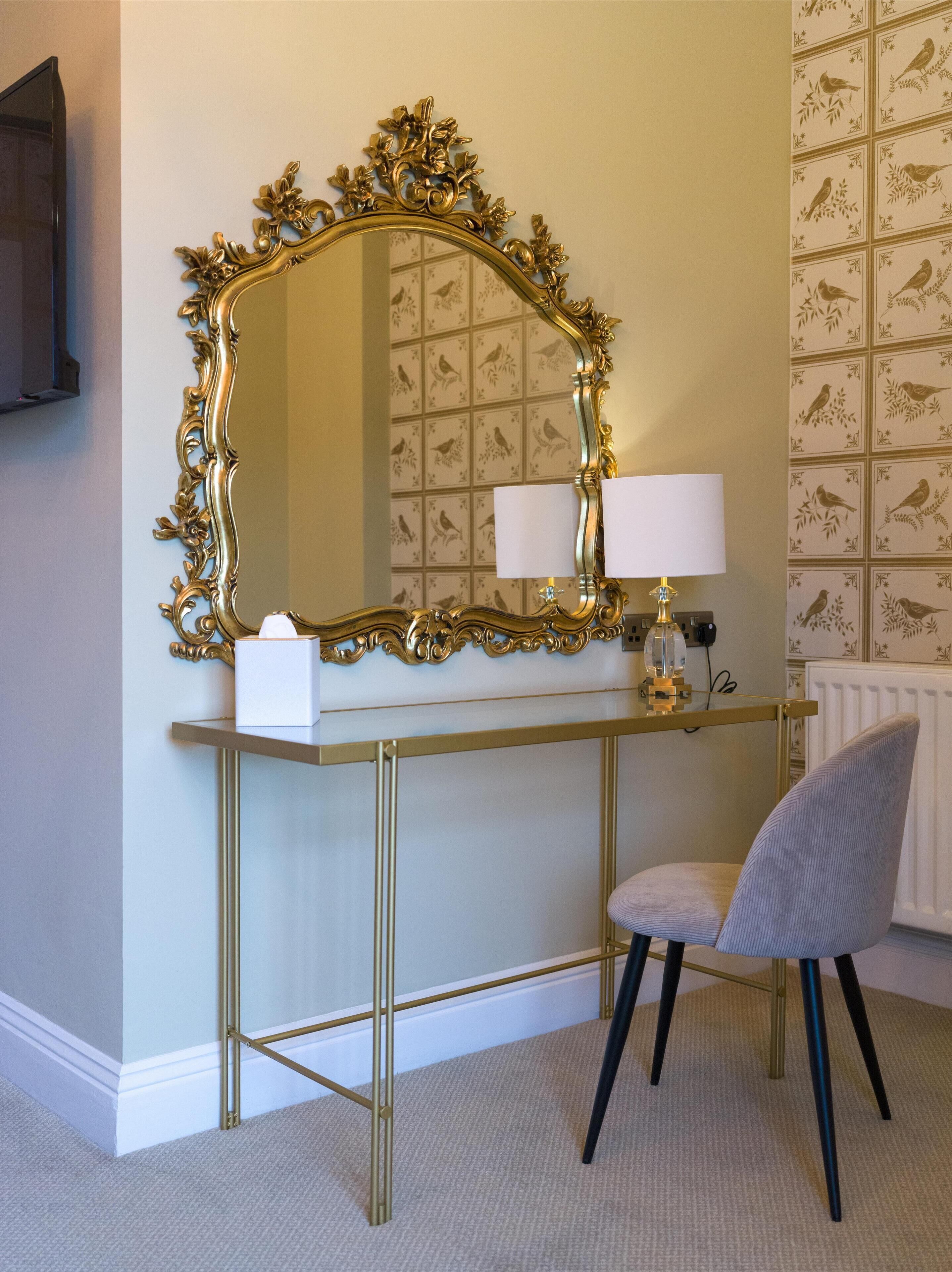 A room with a gold-framed mirror, a small table with a lamp, and a chair.
