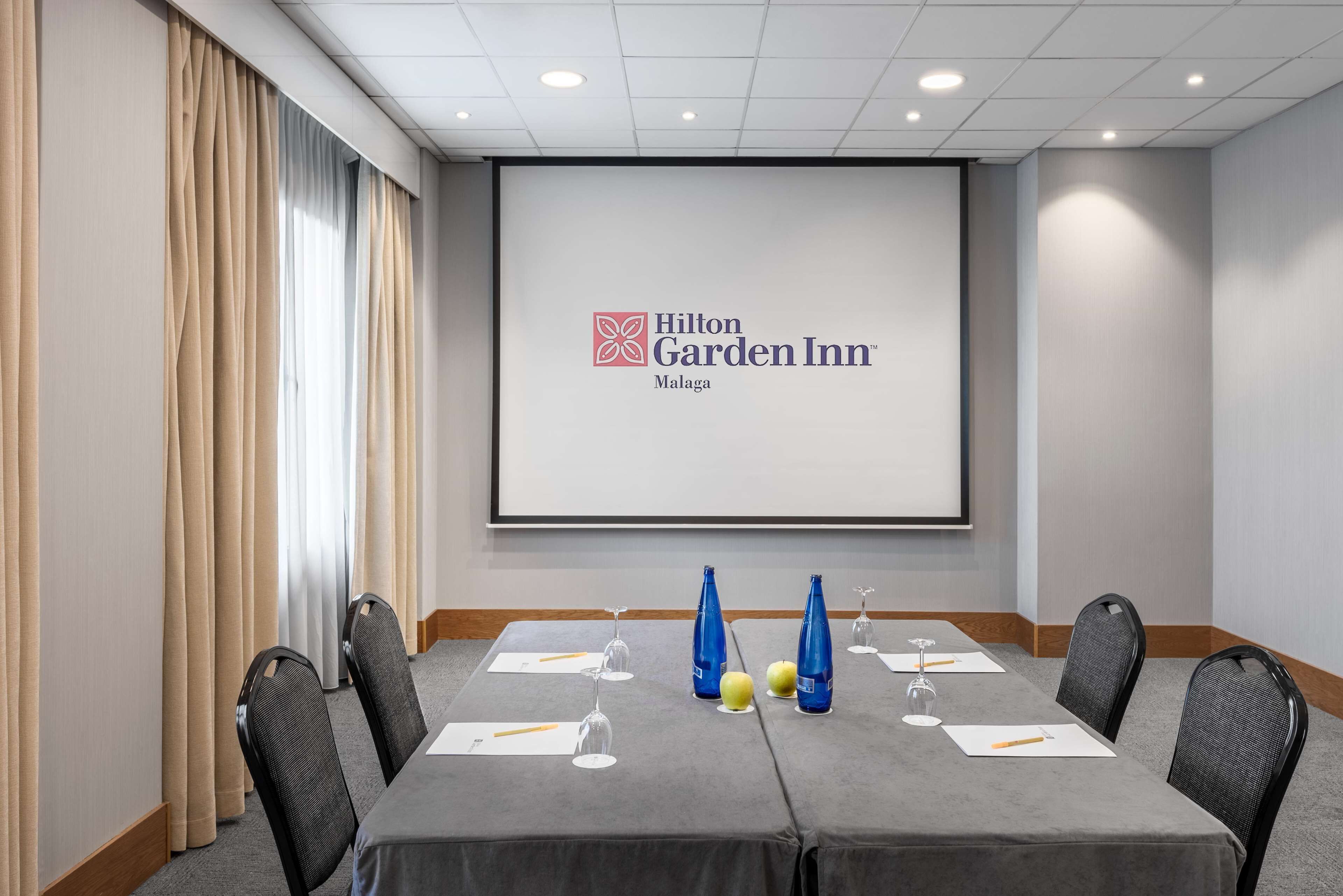 Et mødelokale på Hilton Garden Inn Malaga med et stort skærmbillede af hotellets logo, et langt bord med stole og vandflasker.