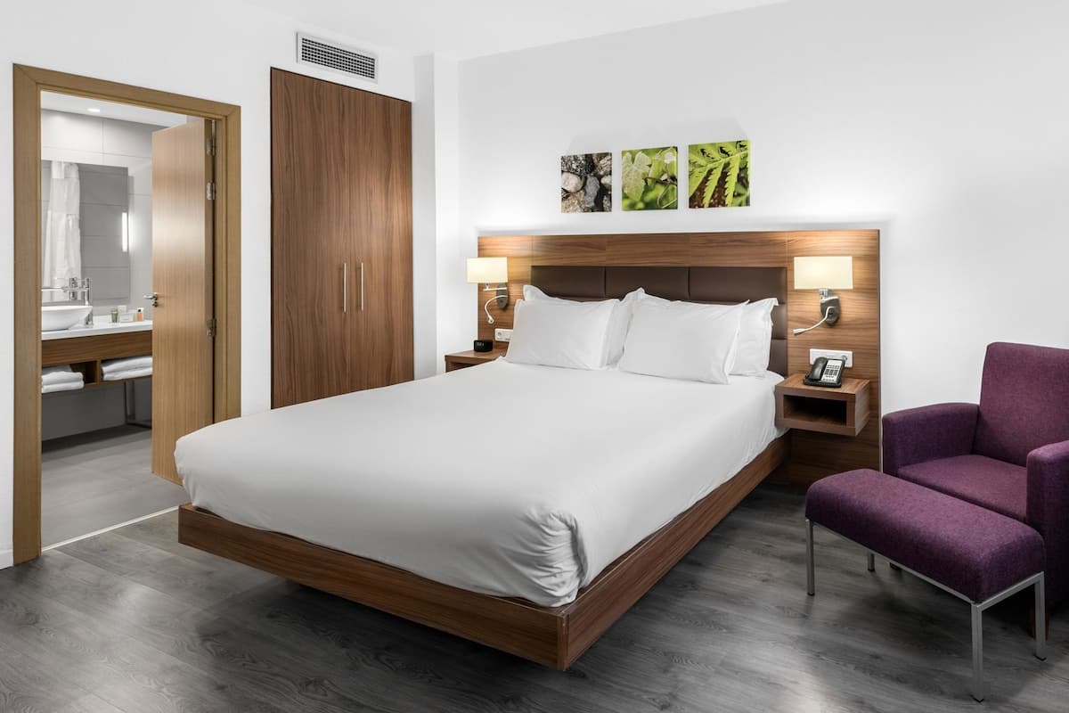 A hotel room with a large bed, a wooden wardrobe, a bathroom with a mirror, and a purple armchair.