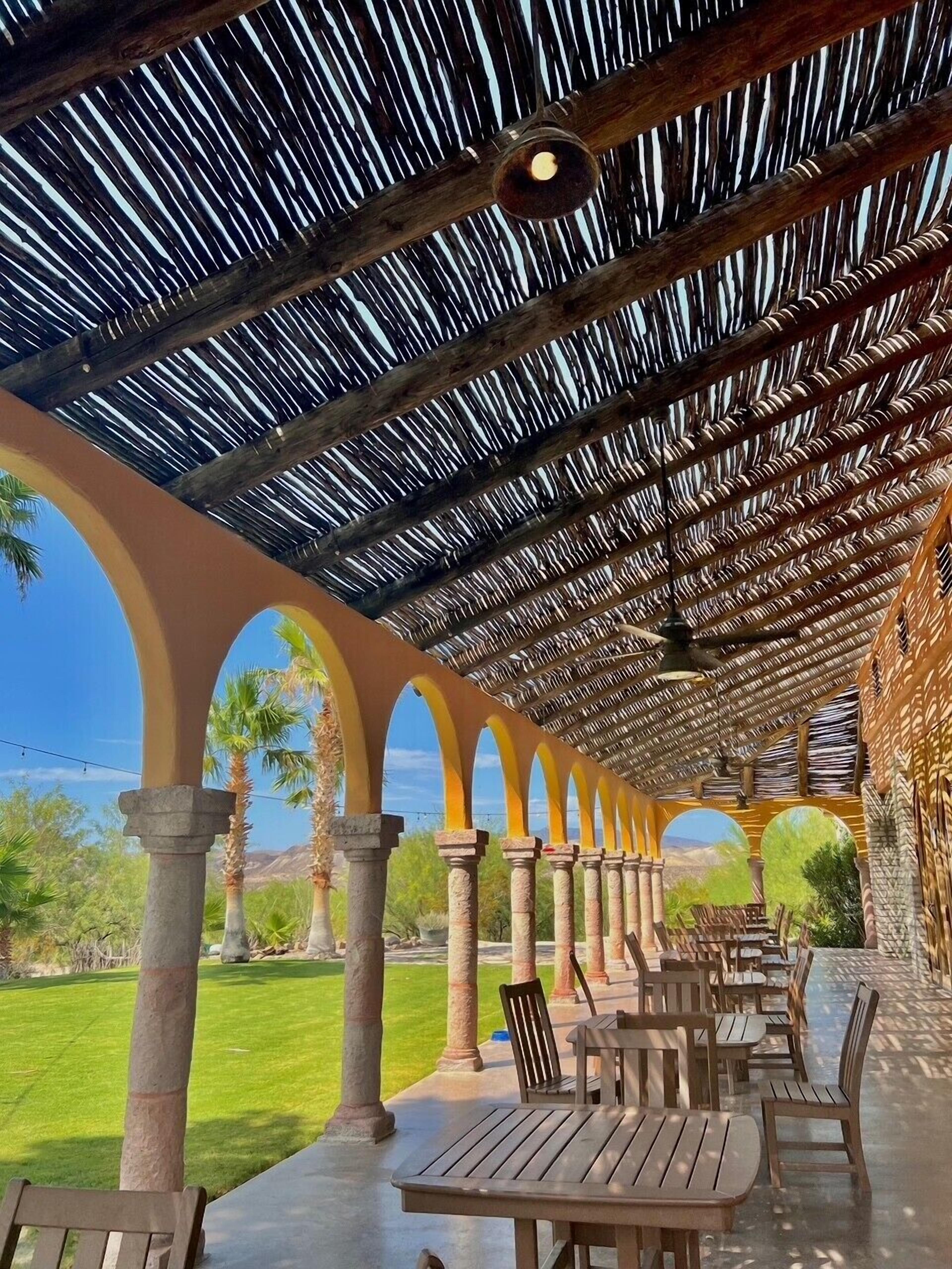 Un patio al aire libre con mesas y sillas de madera, techo de paja y vistas del paisaje.