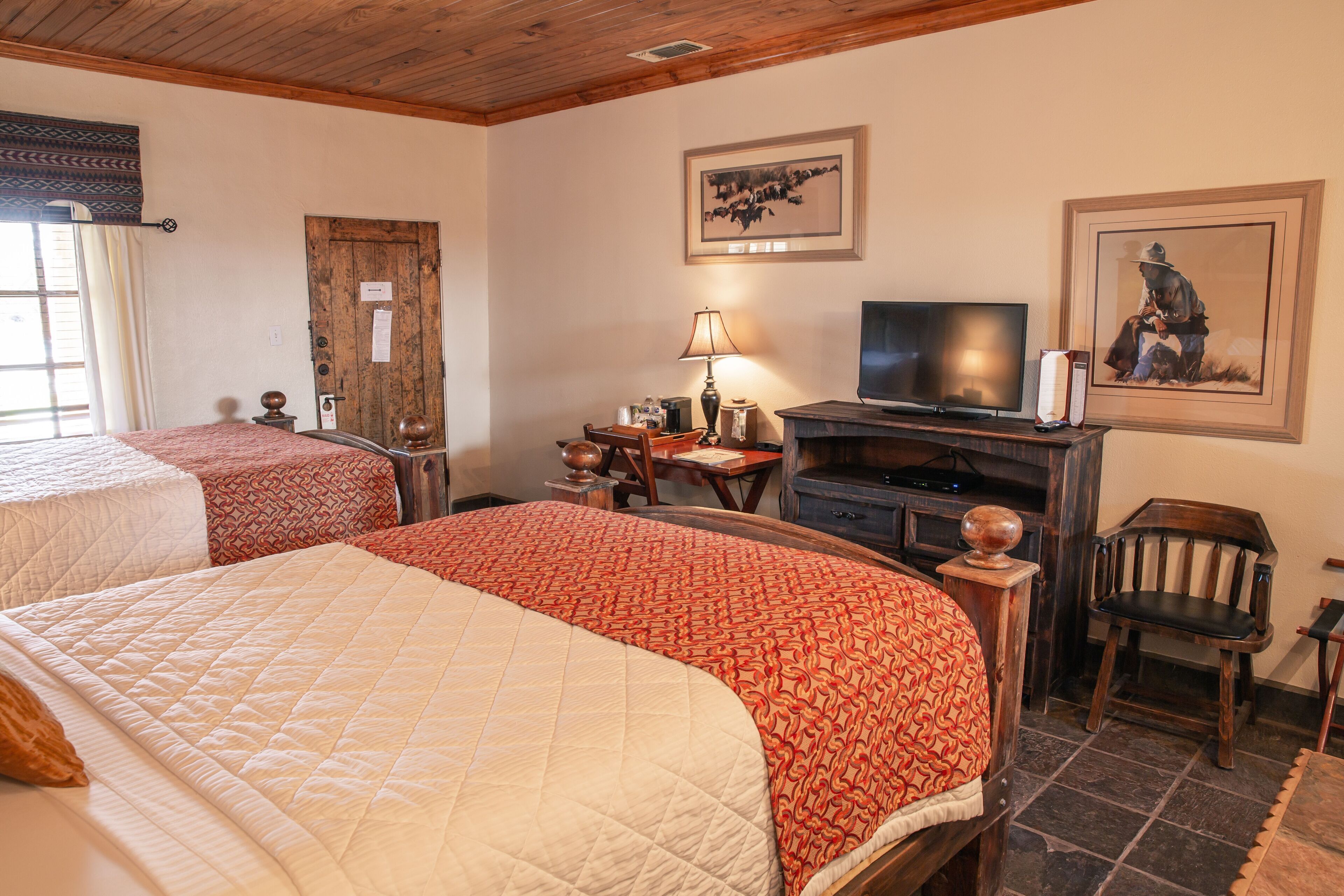 A hotel room with two beds, a television, a desk with a lamp, and artwork on the walls.