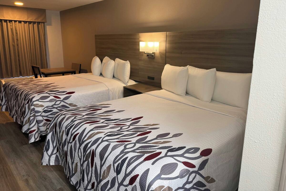 A hotel room with two beds, a desk, a chair, and a wall-mounted lamp.